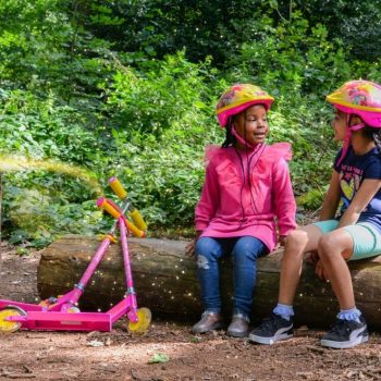 two little girls sitting on a wooden log with pink push scooters next to them